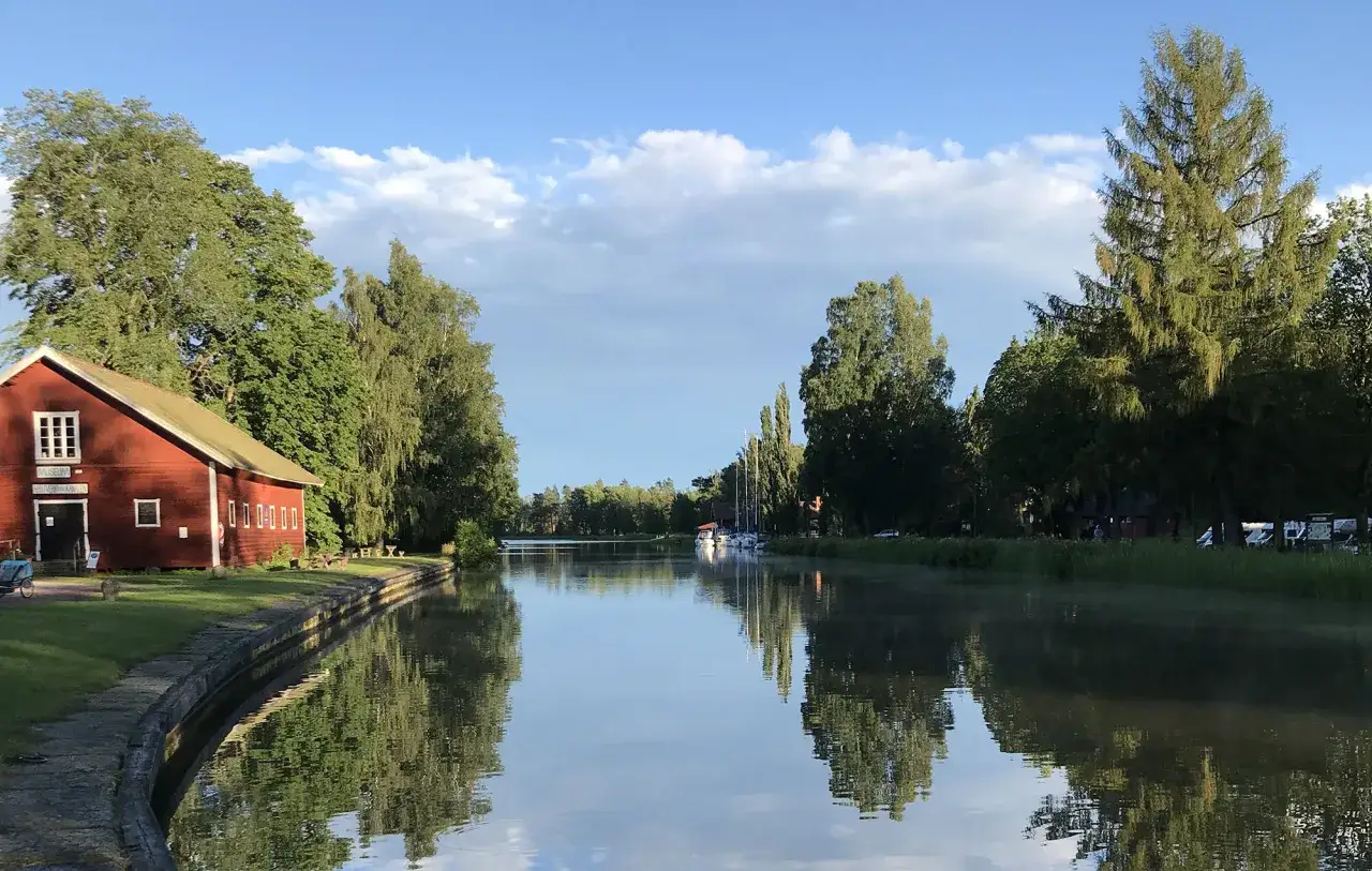 R&ouml;tt hus vid en kanal, en idyllisk plats som p&aring;minner om Hajstorp vandrarhem. Vattnet speglar tr&auml;d och himmel.