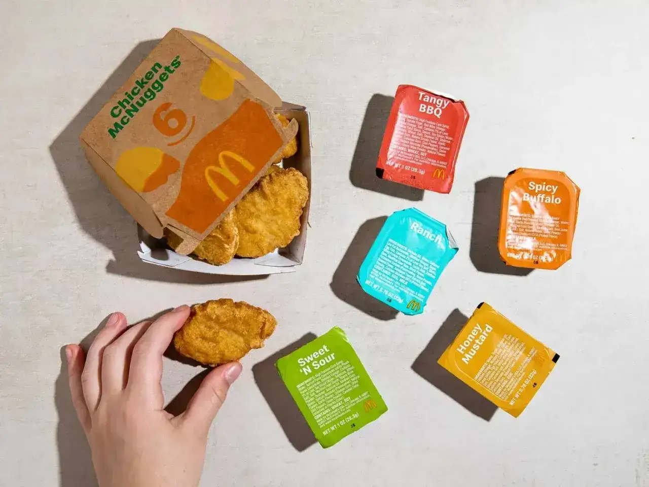 Ręka sięga po McNuggeta z pudełka, obok sosy: Tangy BBQ, Spicy Buffalo, Ranch, Sweet 'N Sour i Honey Mustard. Skład nuggetsy McDonald to tajemnica, ale sosy są dostępne.