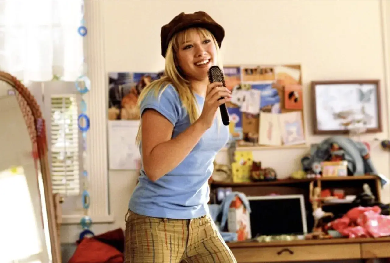 Hilary Duff singt mit einer Haarbürste in ihrem Zimmer, eine Szene, die an ihre Filme und Serien erinnert.