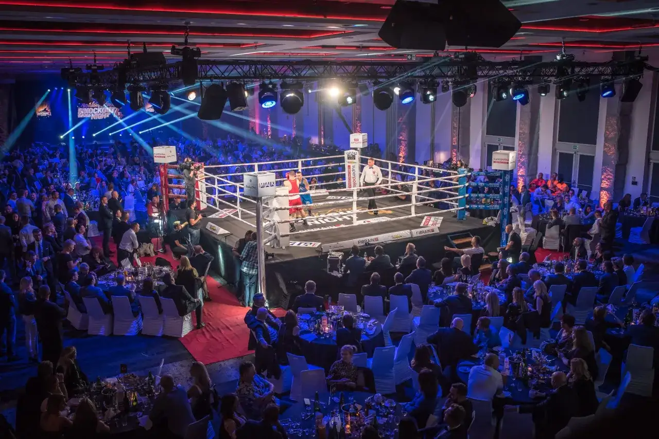 Widownia w eleganckich strojach obserwuje zmagania bokserów podczas **rocky boxing night**.
