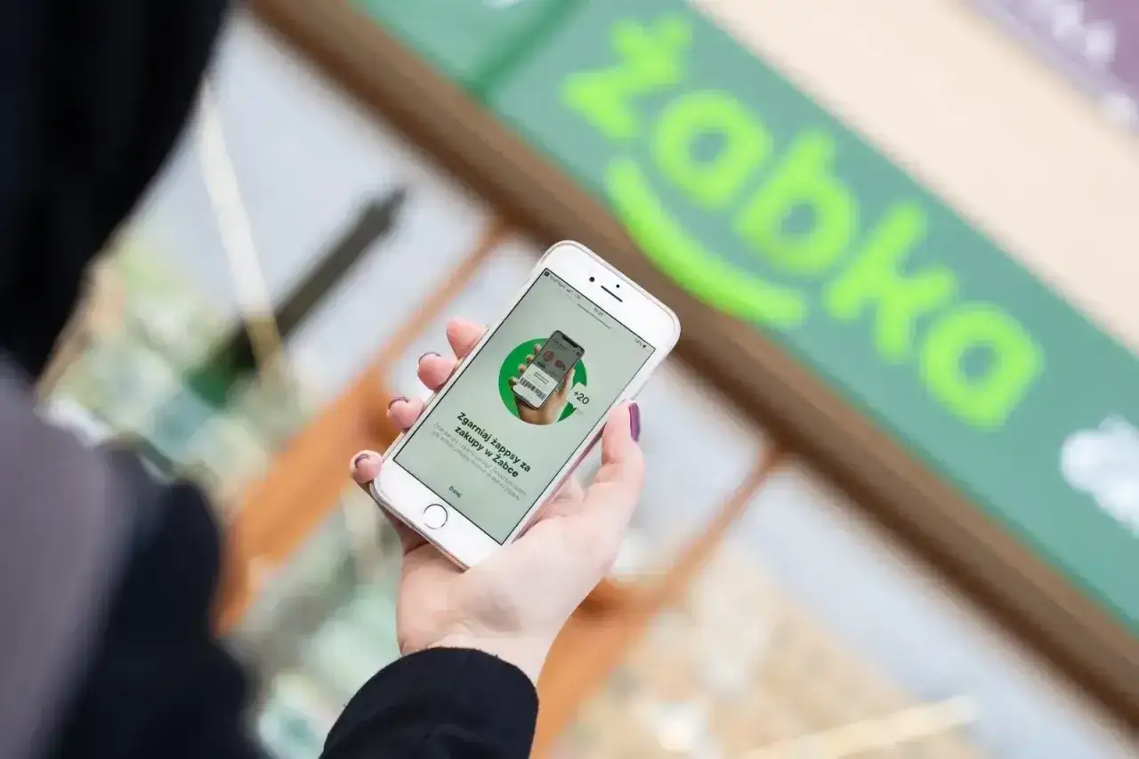Dłoń trzyma smartfon z otwartą aplikacją Żappka, która zachęca do zbierania żappsów za zakupy.