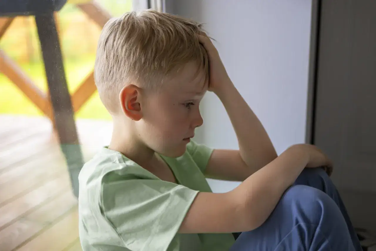 Un niño rubio, con la mano en la cabeza, mira hacia abajo con tristeza. Su postura y expresión sugieren depresión infantil.