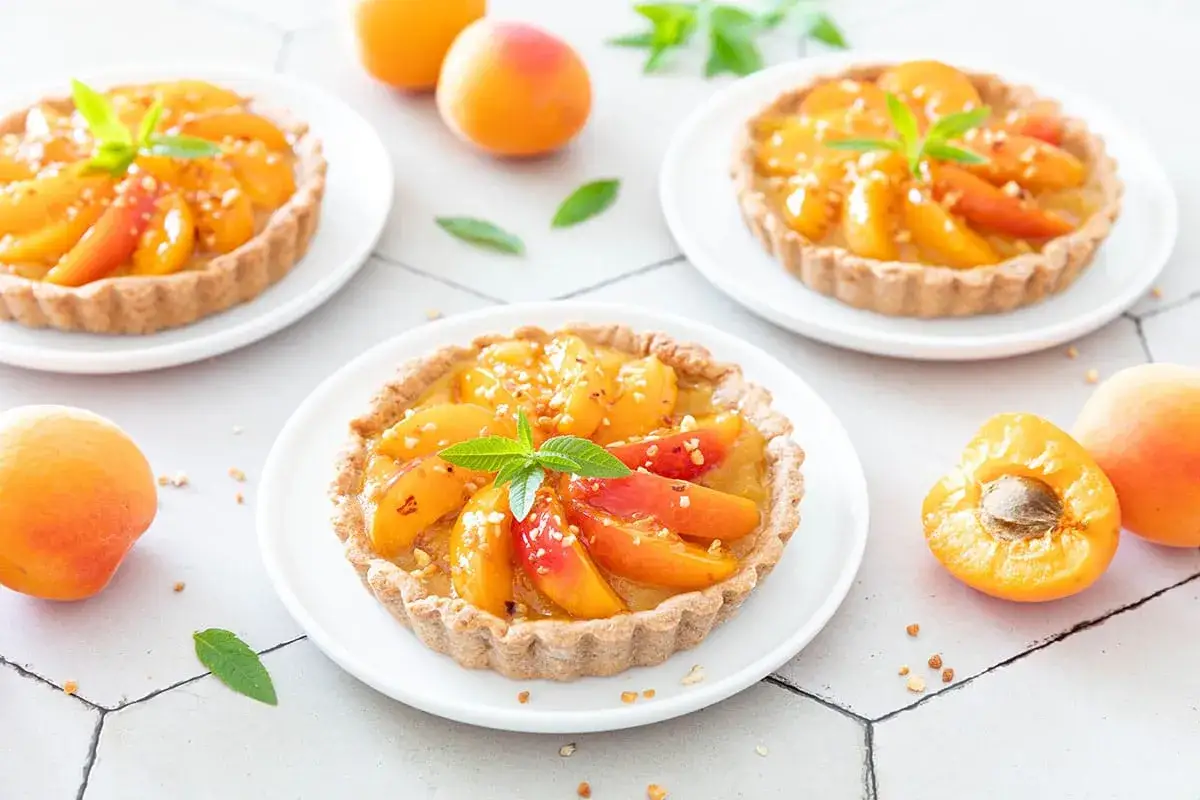 Trois tartelettes &agrave; l'abricot, garnies de fruits frais et de menthe, sur des assiettes blanches. Des abricots entiers et coup&eacute;s sont dispos&eacute;s autour.