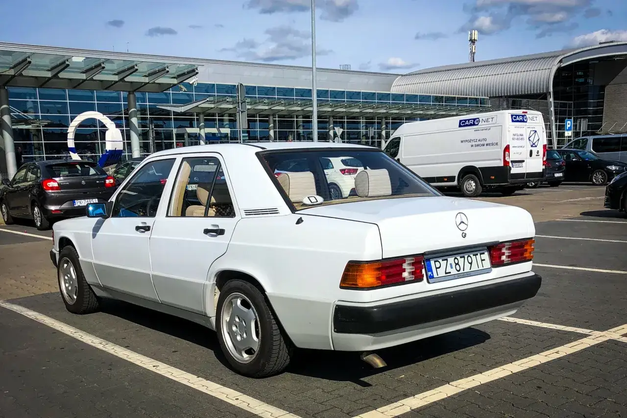 Biały Mercedes 190e na parkingu przed nowoczesnym budynkiem. Klasyk w świetnym stanie.