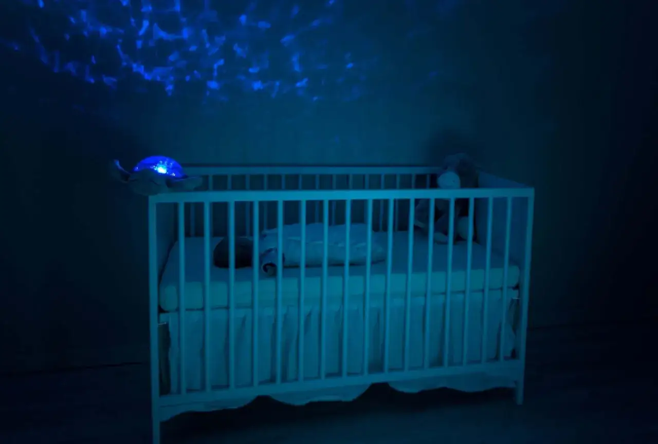 Ein Baby schl&auml;ft in einem Gitterbett, w&auml;hrend ein Nachtlicht mit Sternenprojektion den Raum in blaues Licht taucht. Vielleicht hilft es gegen die schlafregression baby.