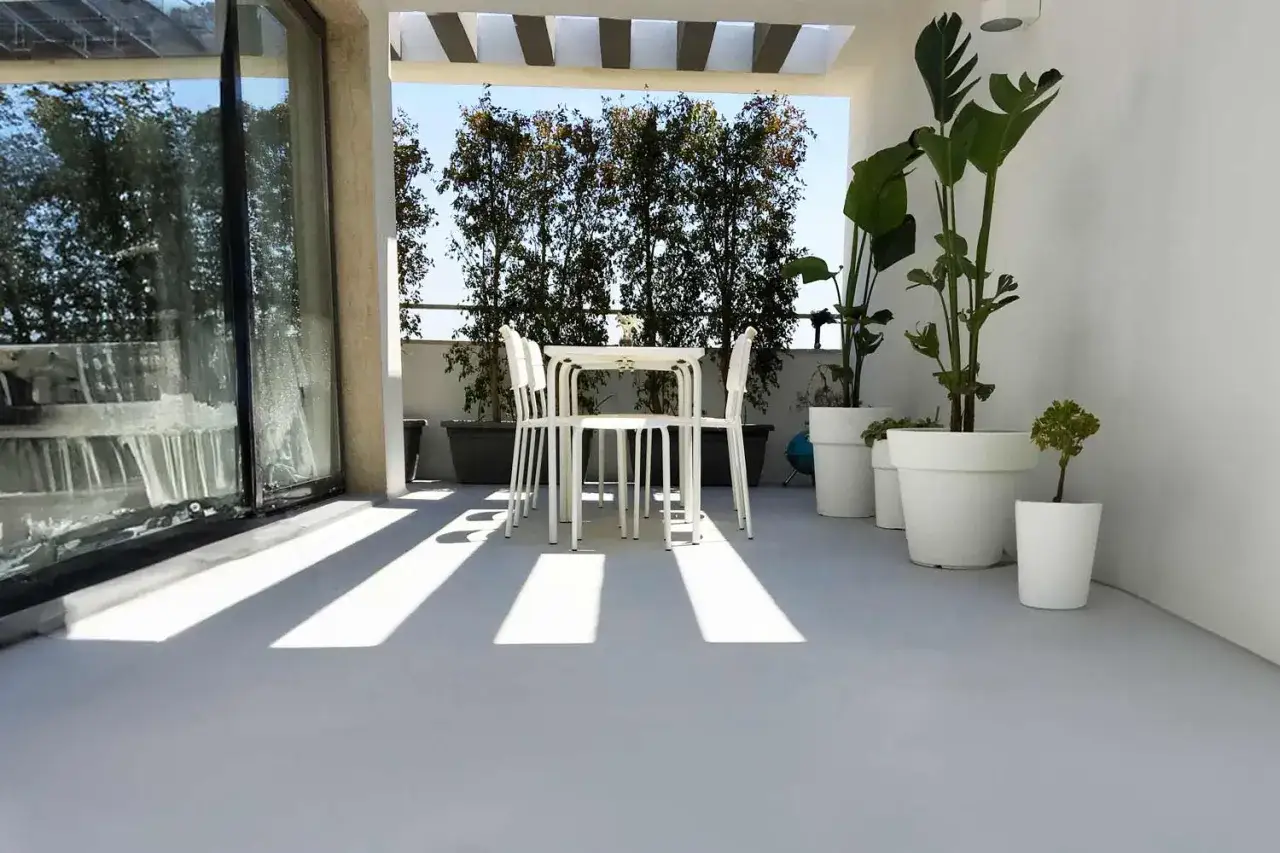 Terrazza moderna con pavimenti in resina per esterni. Tavolo e sedie bianchi, piante in vaso e un'ampia vetrata.