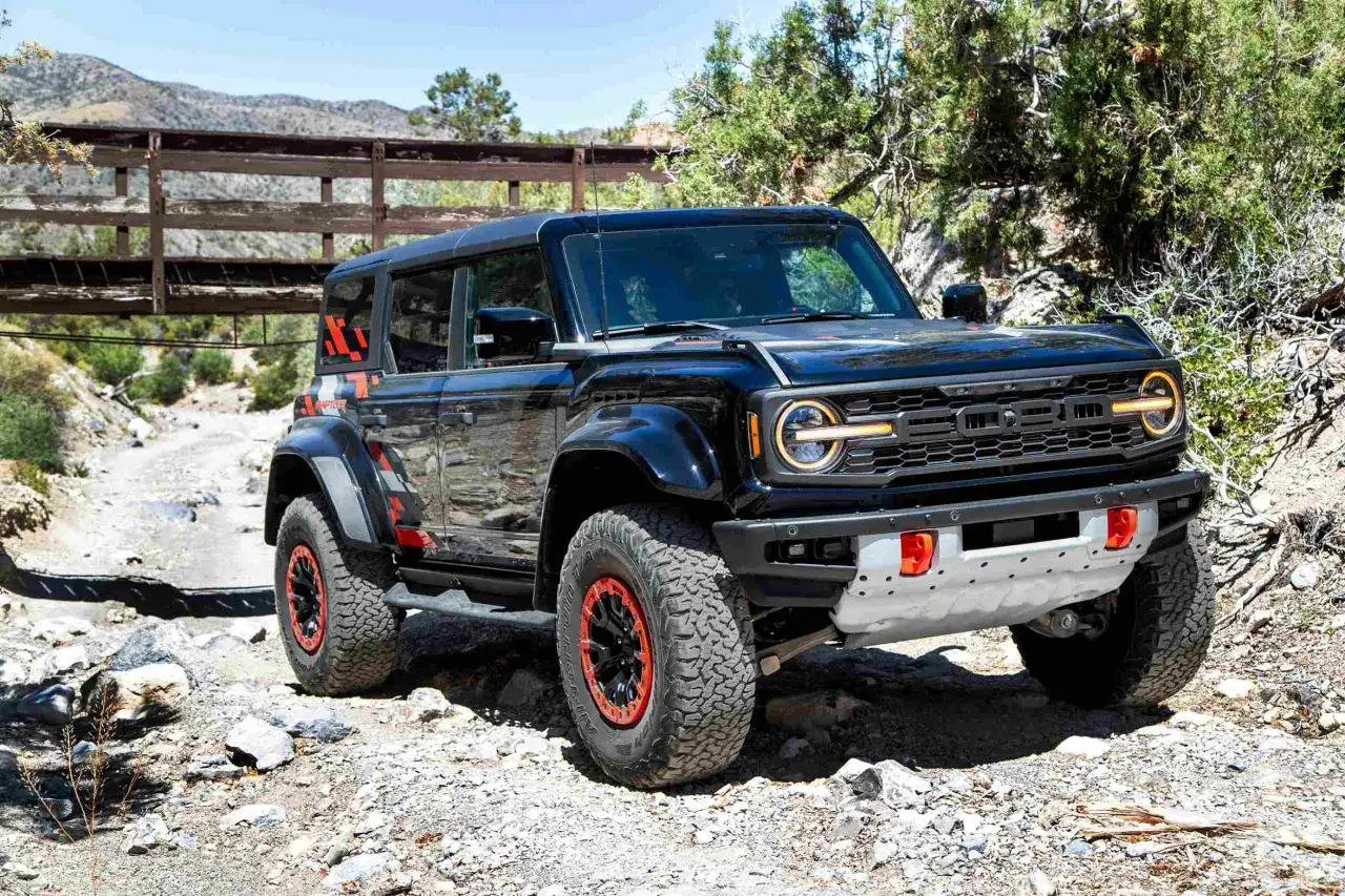 Czarny Ford Bronco Raptor z pomarańczowymi akcentami na felgach i karoserii, gotowy do jazdy po kamienistym, pustynnym terenie pod drewnianym mostkiem.
