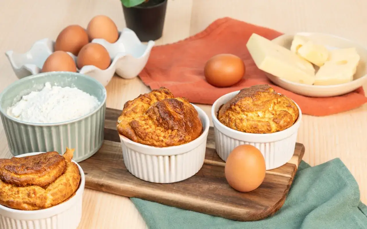 Trois souffl&eacute;s au fromage dor&eacute;s et gonfl&eacute;s dans des ramequins blancs, entour&eacute;s d'&oelig;ufs, de farine et de fromage.