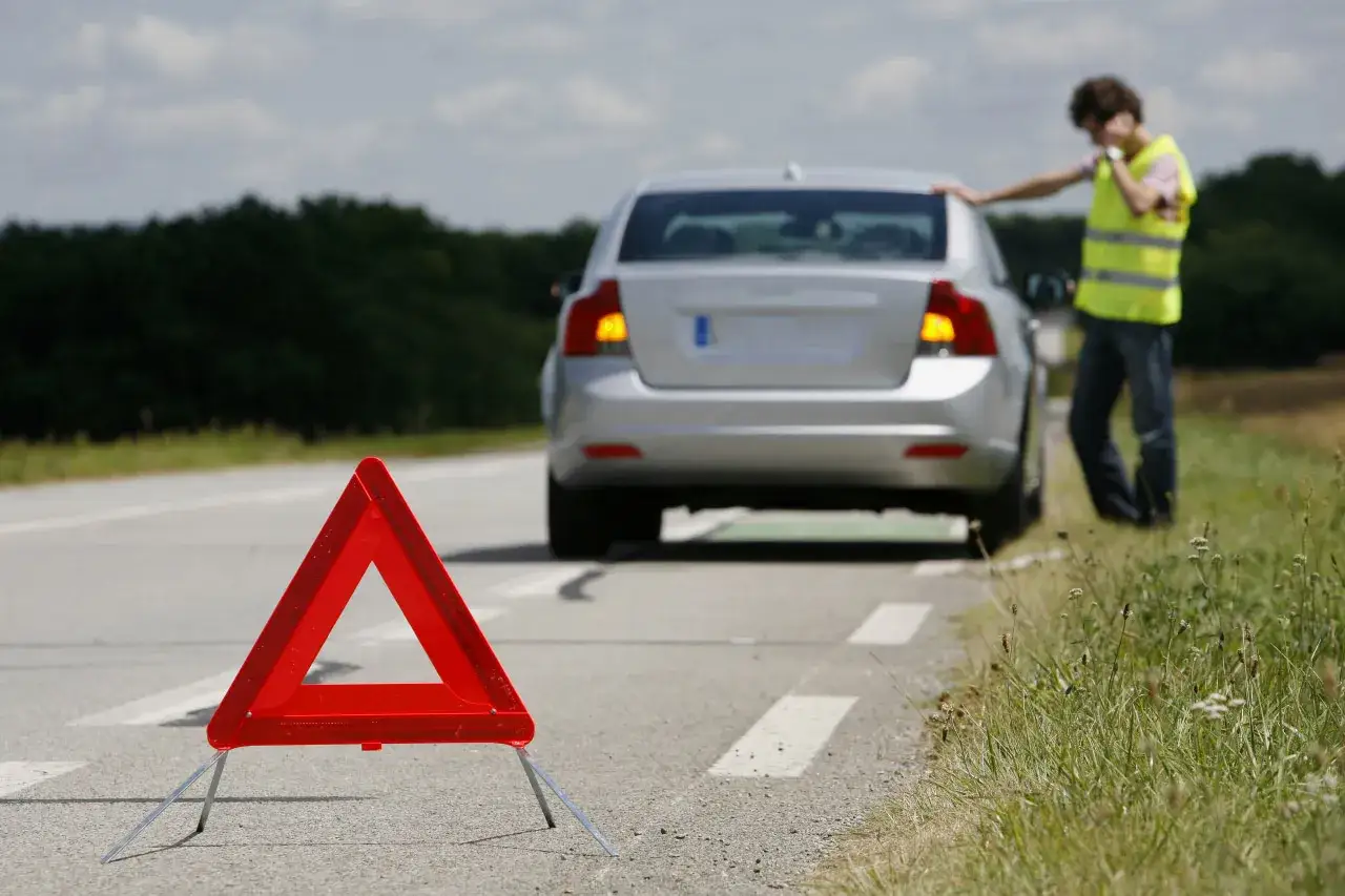 Tri&aacute;ngulo de emergencia rojo en la carretera. Un hombre con chaleco reflectante habla por tel&eacute;fono junto a un coche plateado averiado. &iquest;Qu&eacute; es race?