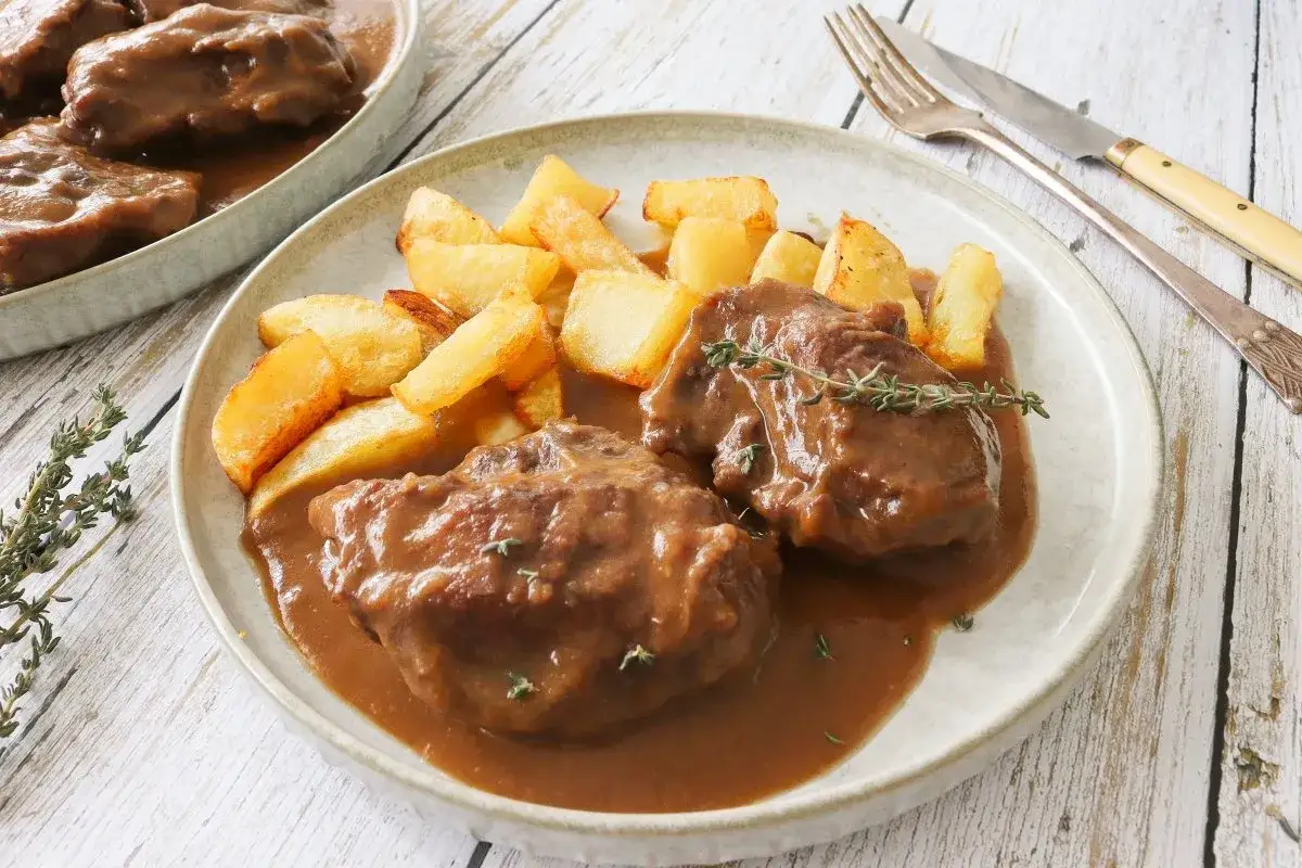 Dos tiernas patas de ternera guisada en su jugo, acompañadas de patatas rústicas fritas y una ramita de tomillo fresco.