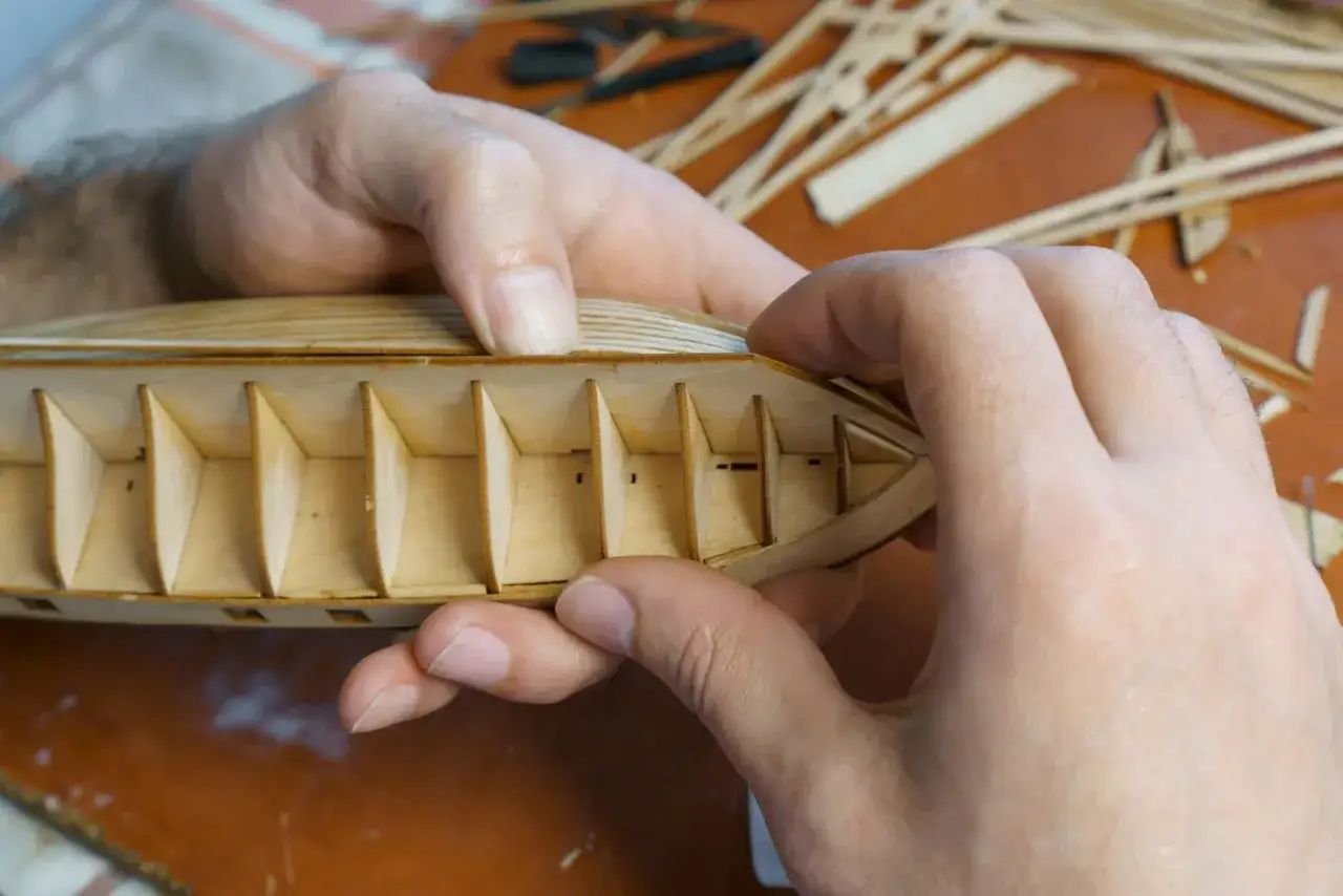 Hände bauen einen hölzernen Schiffs-Kadlub. Kleine Holzspanten sind sichtbar.