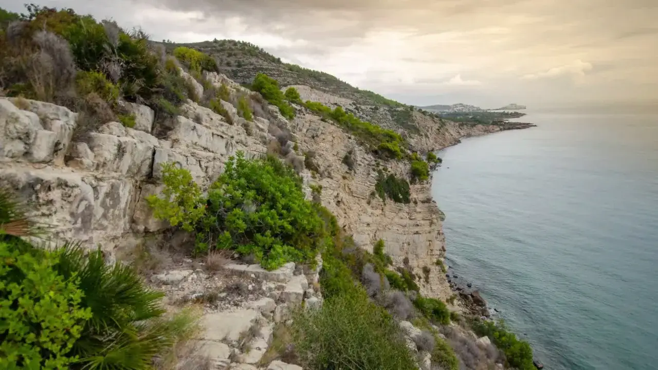 Acantilados rocosos y vegetación agreste en la **sierra de Irta**, con el mar Mediterráneo extendiéndose hacia el horizonte bajo un cielo nublado.