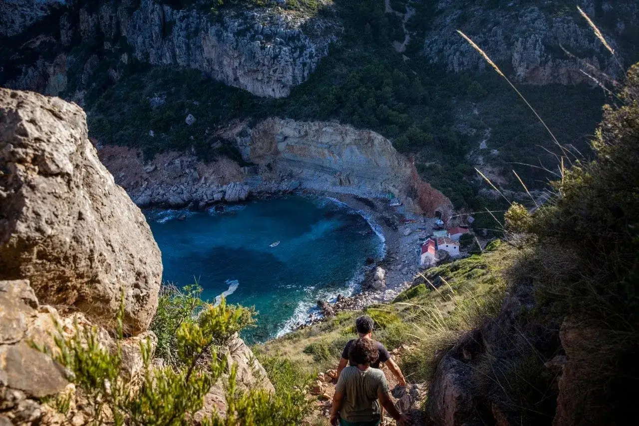 Dos excursionistas descienden por un sendero rocoso hacia la **cala dels testos**, una bahía de aguas turquesas con casitas blancas y un pequeño bote.