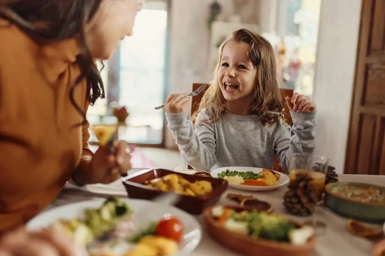 Bambina con cibo in bocca, sorride mentre mangia con la famiglia.
