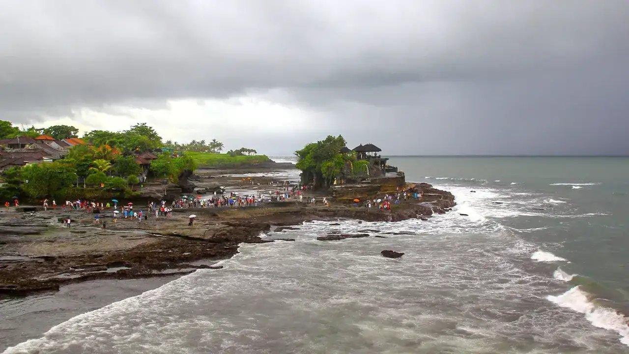 Tłumy ludzi zwiedzają świątynię Tanah Lot na Bali podczas pory deszczowej. Chmury zapowiadają deszcz, ale fale oceanu wciąż rozbijają się o skaliste wybrzeże.