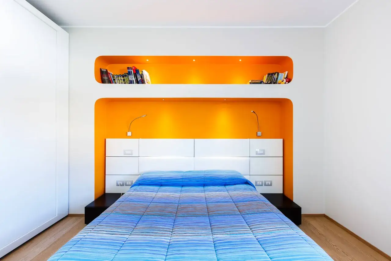Cameretta moderna con nicchie arancioni in cartongesso che fungono da libreria, letto bianco e copriletto a righe blu.
