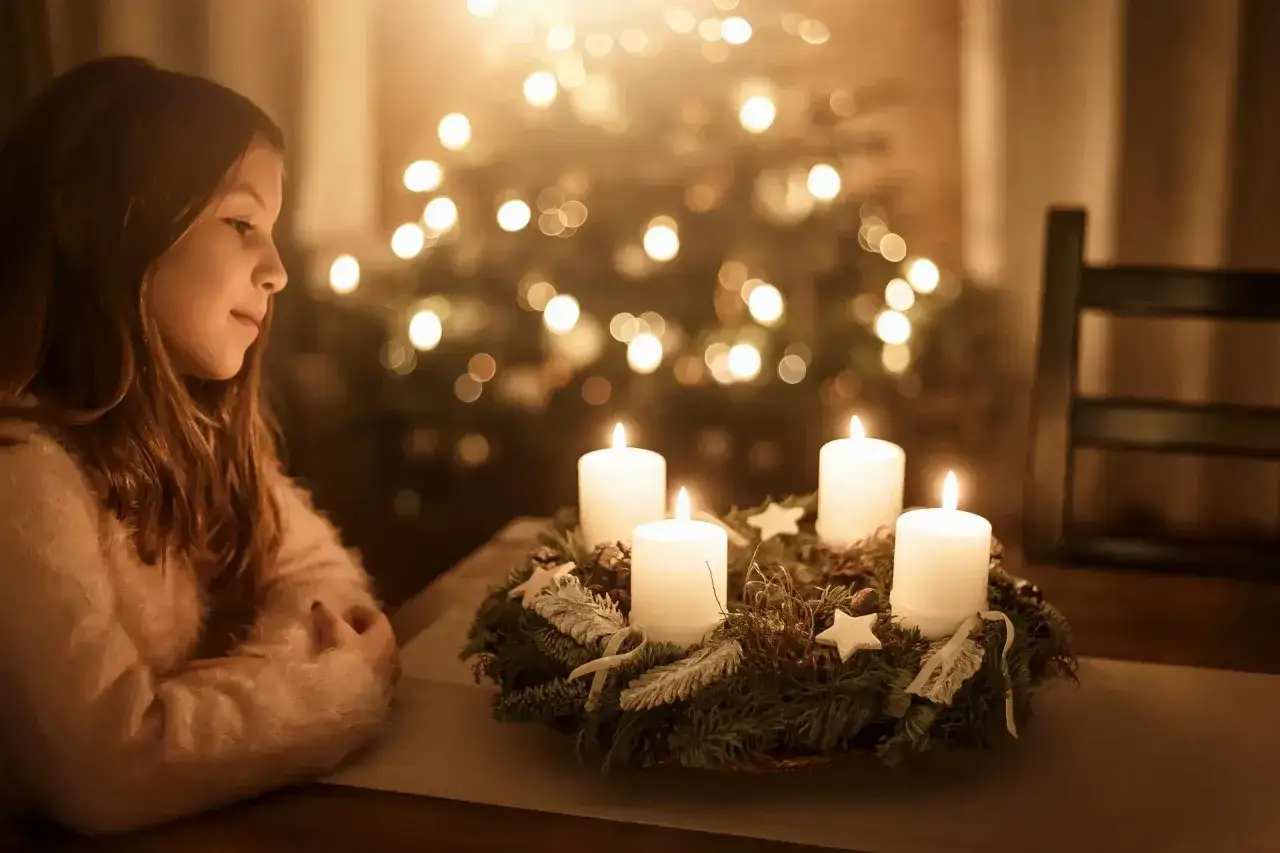 Ein M&auml;dchen blickt auf einen Adventskranz mit vier brennenden Kerzen. Im Hintergrund leuchtet ein Weihnachtsbaum. Eine besinnliche Weihnachtsstimmung.