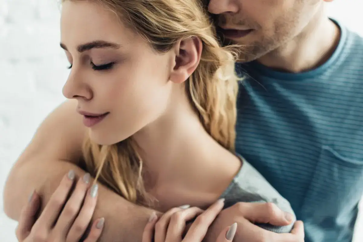Blondynka z zamkniętymi oczami przytulona przez mężczyznę. Jej dłonie spoczywają na jego ramieniu, jakby walczyła z filofobią, szukając ukojenia.