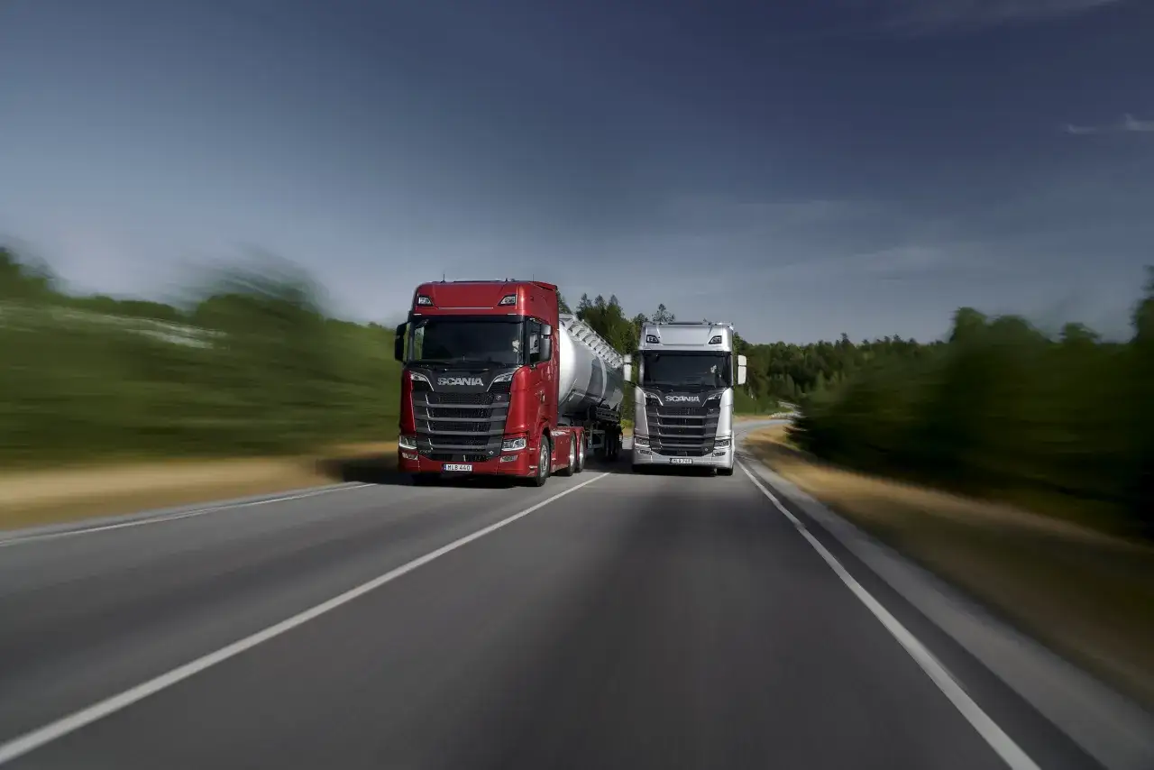 Dwa potężne ciągniki Scania V8, jeden czerwony, drugi srebrny, pędzą po autostradzie wśród zieleni.