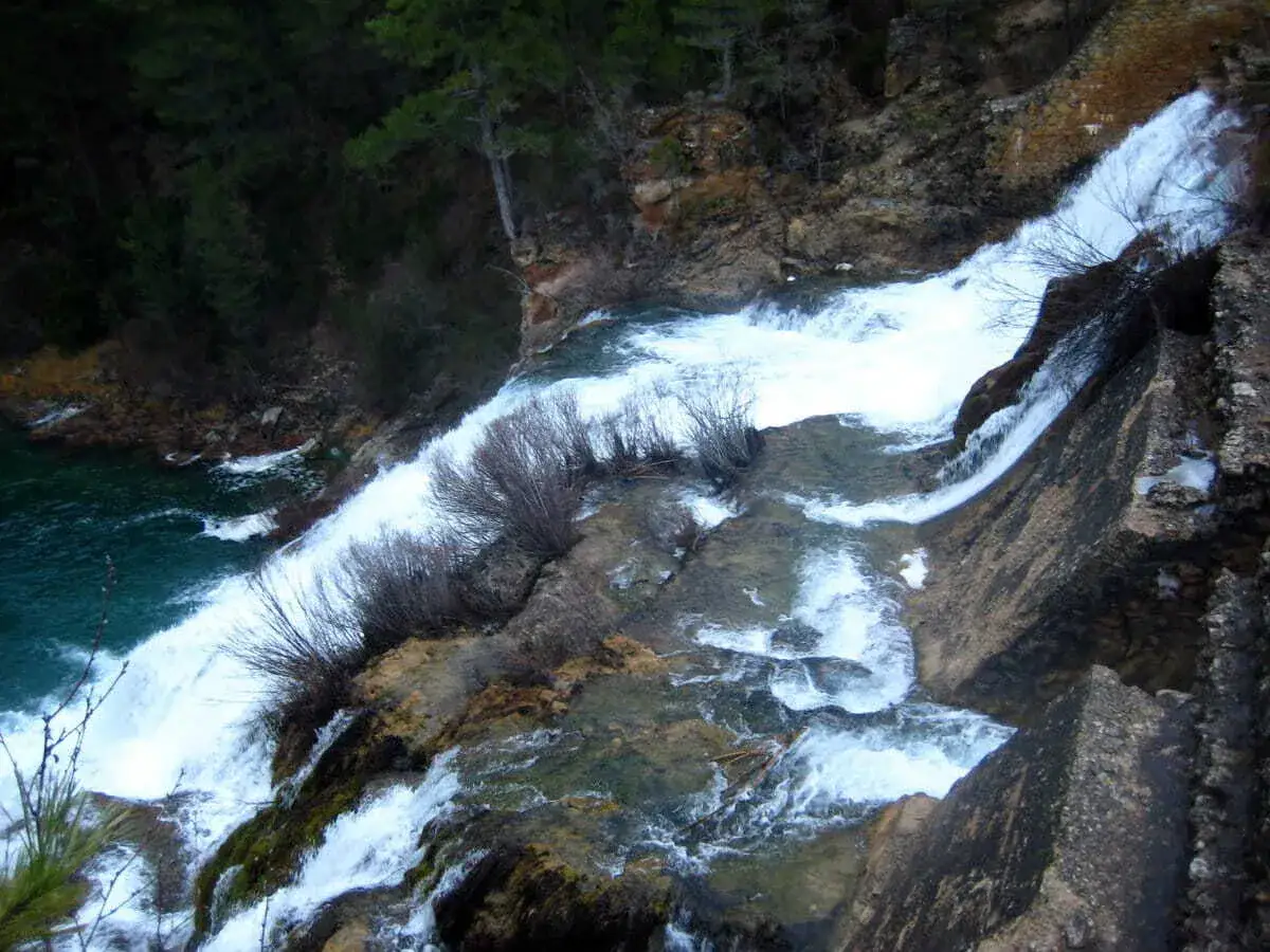 El salto de Poveda, una cascada de agua blanca y espumosa que desciende por rocas y vegetaci&oacute;n, desemboca en un r&iacute;o de aguas turquesas.