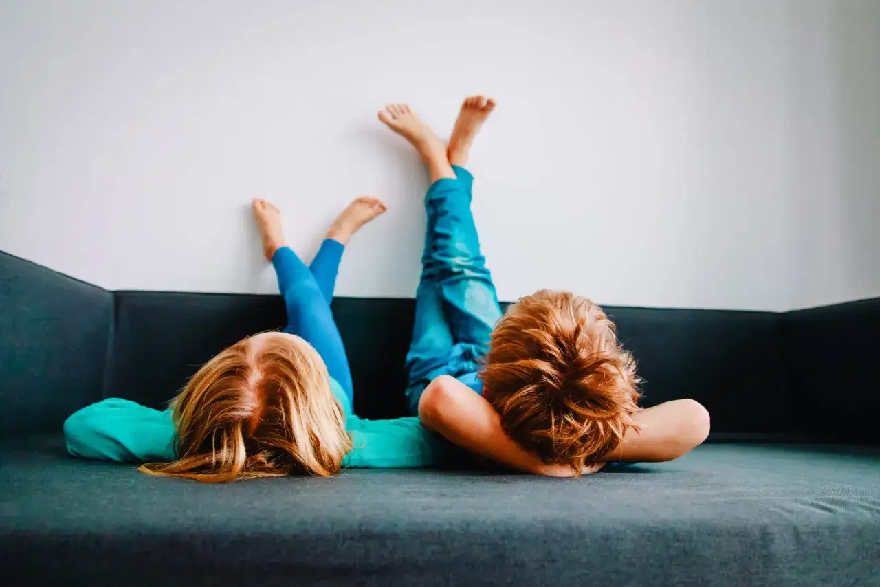 Zwei Kinder liegen kopf&uuml;ber auf einem Sofa, die Beine an der Wand. Sie machen keine Hausaufgaben, sondern entspannen sich.