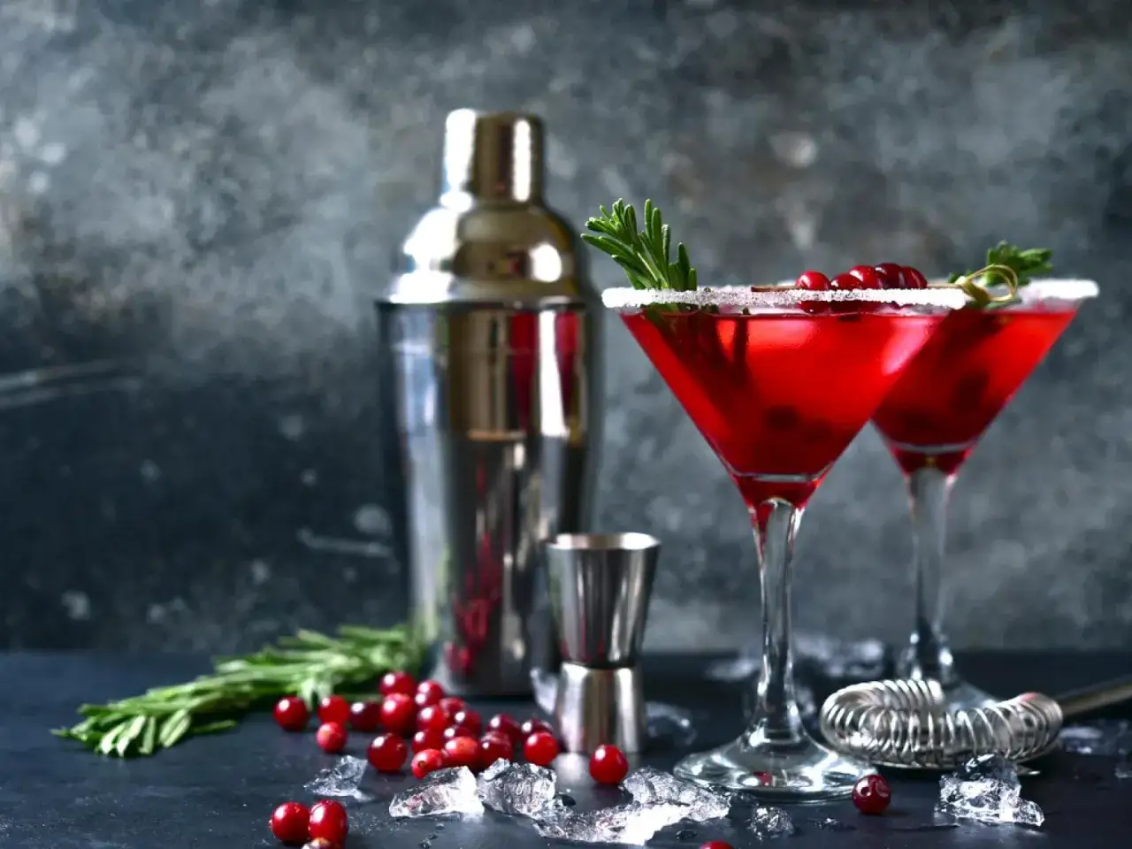 Zwei rote Cocktails im Martini-Glas, garniert mit Rosmarin und Cranberries, neben einem Shaker und Eisw&uuml;rfeln. Ein festlicher Snow Globe Cocktail.
