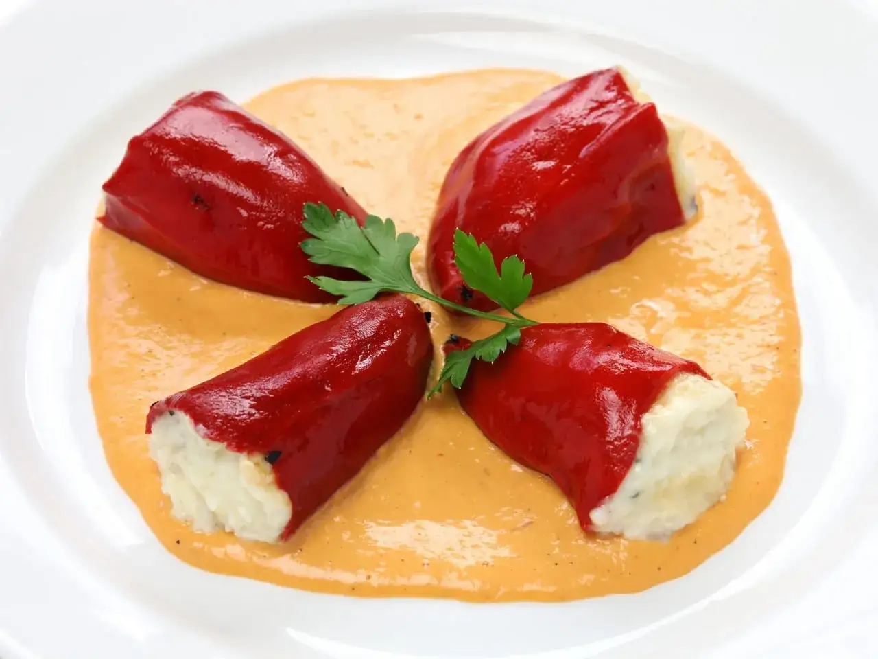 Cuatro pimientos del piquillo rellenos de una pasta blanca, servidos sobre una salsa cremosa y decorados con perejil.