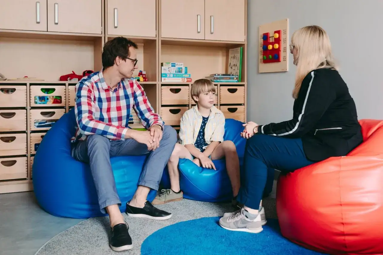 Rodzina rozmawia z terapeutą o objawach zespołu Aspergera. Dziecko siedzi na niebieskiej pufie, rodzice na czerwonej i niebieskiej.
