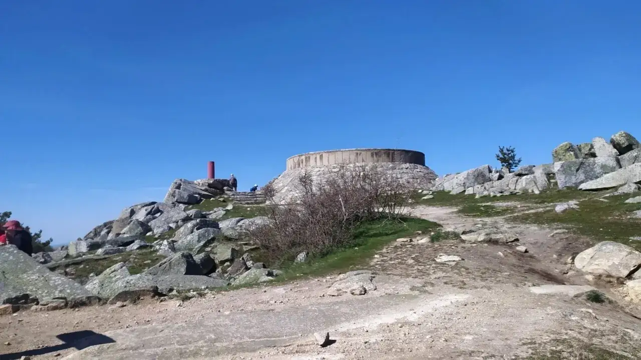 Vista de una estructura circular de hormig&oacute;n y piedra en una colina rocosa, con un poste rojo y personas. Parece una cabeza lijar en la cima.