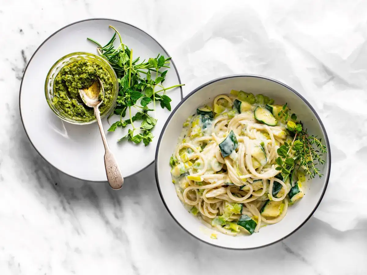 Pyszny makaron z cukinią w kremowym sosie, obok miseczka pesto i świeże zioła.