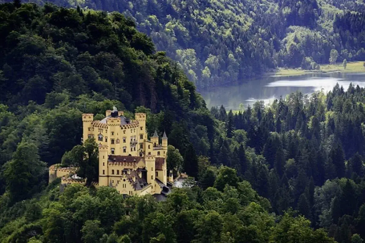 Gelbes Schloss thront auf einem H&uuml;gel inmitten dichter W&auml;lder, mit einem See im Hintergrund. Ein M&auml;rchenbild, das zu den sch&ouml;nsten Attraktionen Bayerns z&auml;hlt.
