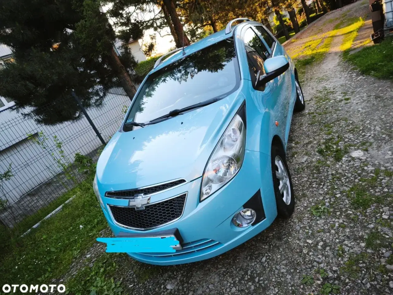 Błękitny Chevrolet Spark 2010 zaparkowany na żwirowej drodze, otoczony zielenią i drzewami.