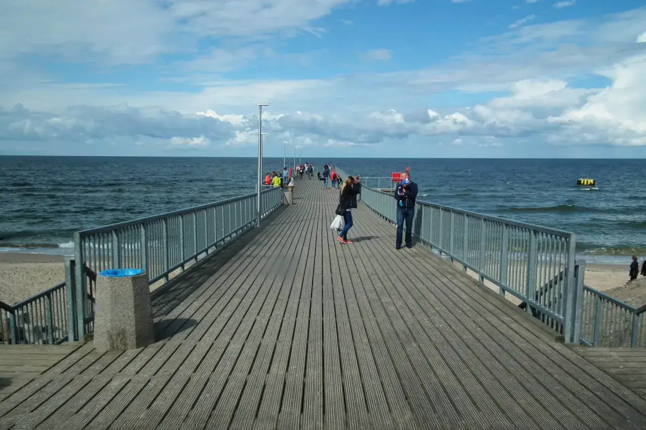 Ludzie spacerują po drewnianym deptaku nad morzem. W oddali widać falujące morze i błękitne niebo z chmurami.