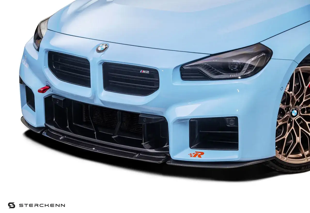 Niebieskie BMW M2 z agresywnym przednim zderzakiem i karbonowym spojlerem. Czerwony uchwyt holowniczy dodaje sportowego charakteru.