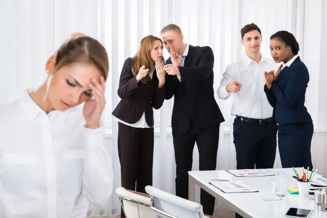 Donna turbata in primo piano, mentre colleghi la deridono. Un esempio di mobbing sul lavoro cosa fare per affrontarlo.