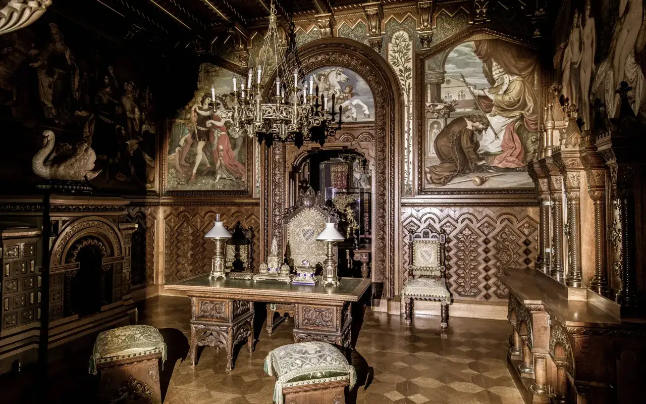 Prunkvoller Schreibtisch mit Lampen und Thronstuhl im Neuschwanstein innen. Wandmalereien und ein Kronleuchter vervollständigen das Bild.