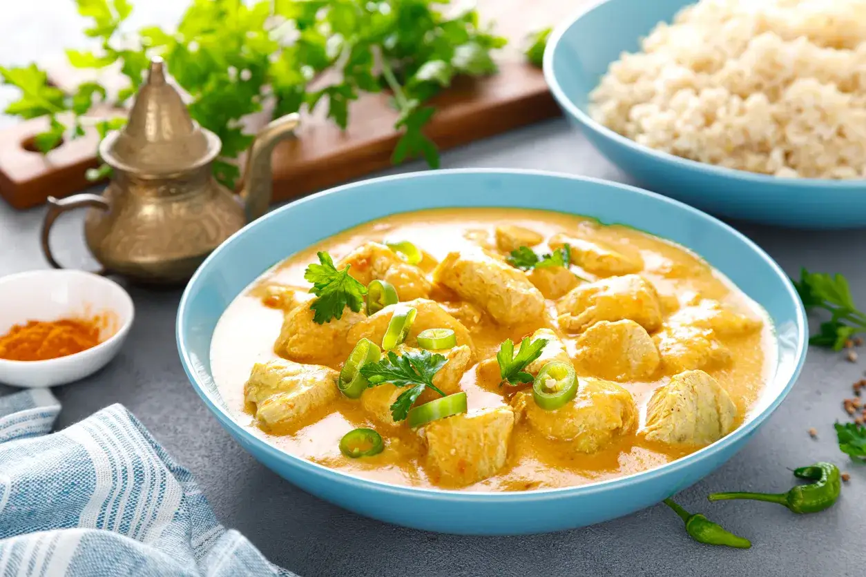 Aksamitny kurczak po tajlandzku w kremowym sosie curry, udekorowany świeżą kolendrą i papryczkami chili. Obok miseczka ryżu i przypraw.