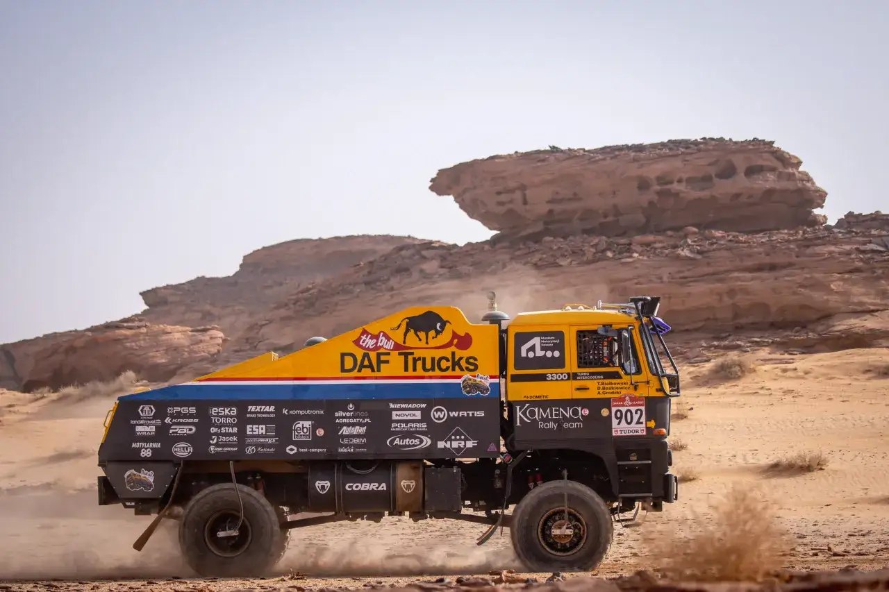 Żółta ciężarówka DAF Trucks z logo Kamena Rally Team pędzi przez pustynię, wzbijając tumany kurzu.
