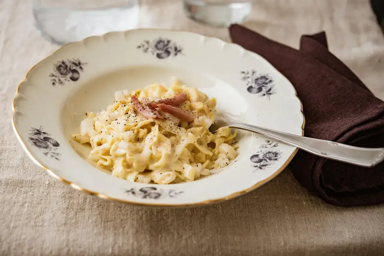 Pyszne tagliatelle w kremowym sosie, posypane pieprzem i kawałkami boczku, podane na ozdobnym talerzu.