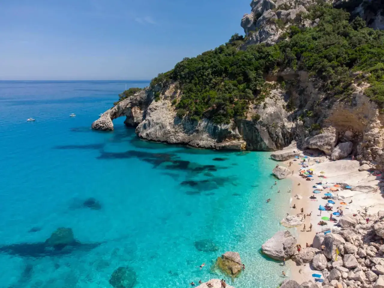 Cala Goloritze, Sardynia. Turkusowa woda, skaliste klify porośnięte zielenią i plaża pełna kolorowych parasoli.