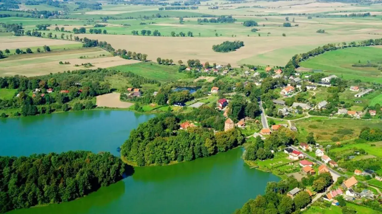 Malownicza wieś nad jeziorem, z zielonymi polami i lasami. W oddali widać przezmark jezioro, otoczone drzewami i domami.