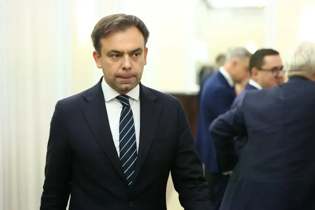 Minister finansów w granatowym garniturze i krawacie, w tle inni mężczyźni w podobnych strojach.
