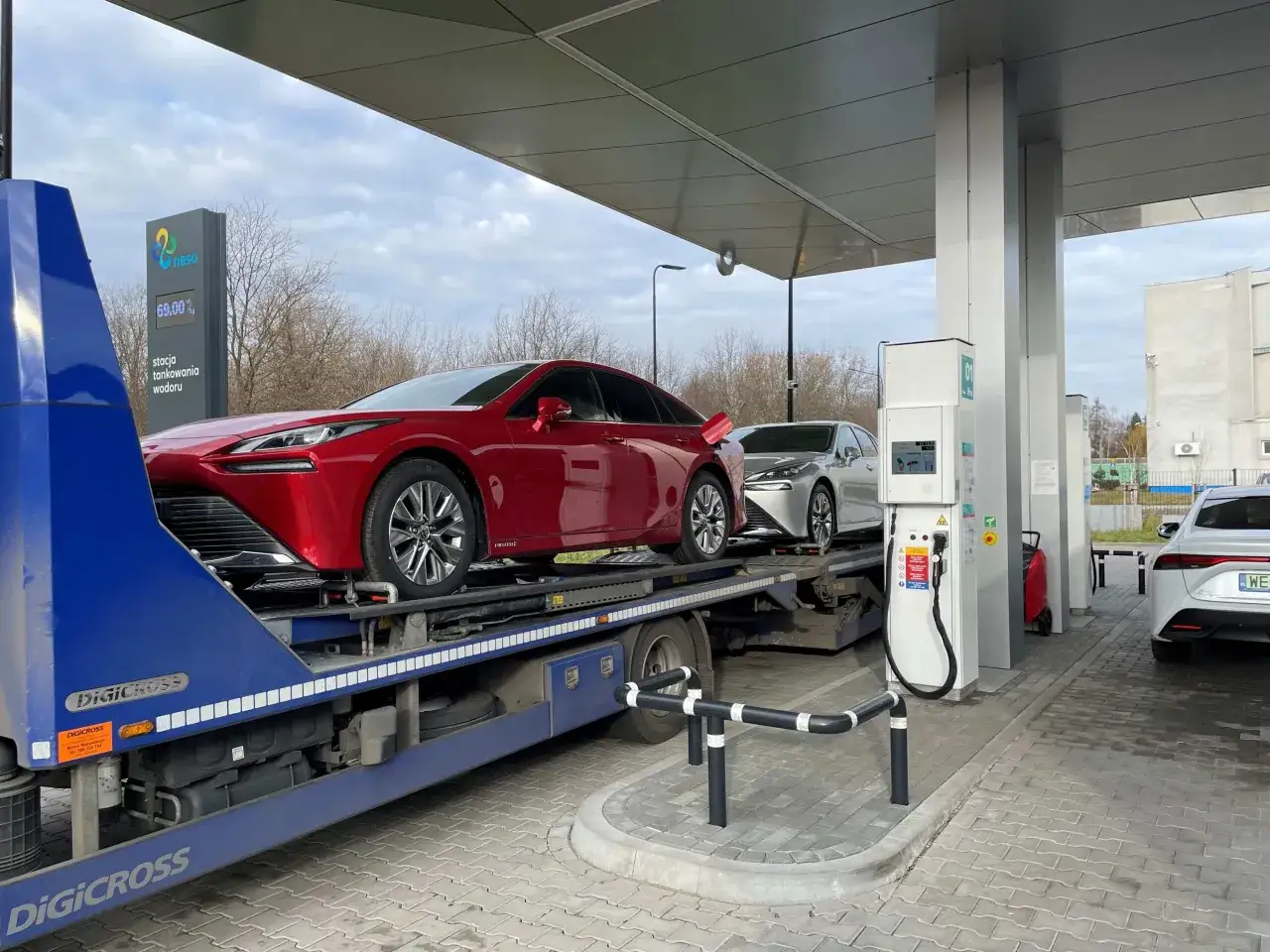 Nowe stacje wodorowe w Polsce to przyszłość motoryzacji. Czerwony i srebrny samochód na platformie transportowej przy dystrybutorze wodoru.
