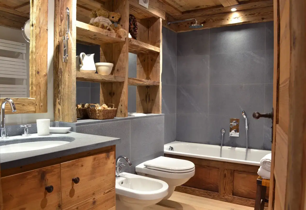 Baños rústicos con madera natural, piedra gris y detalles acogedores. Un lavabo de piedra, inodoro, bidet y bañera crean un espacio funcional y con encanto.