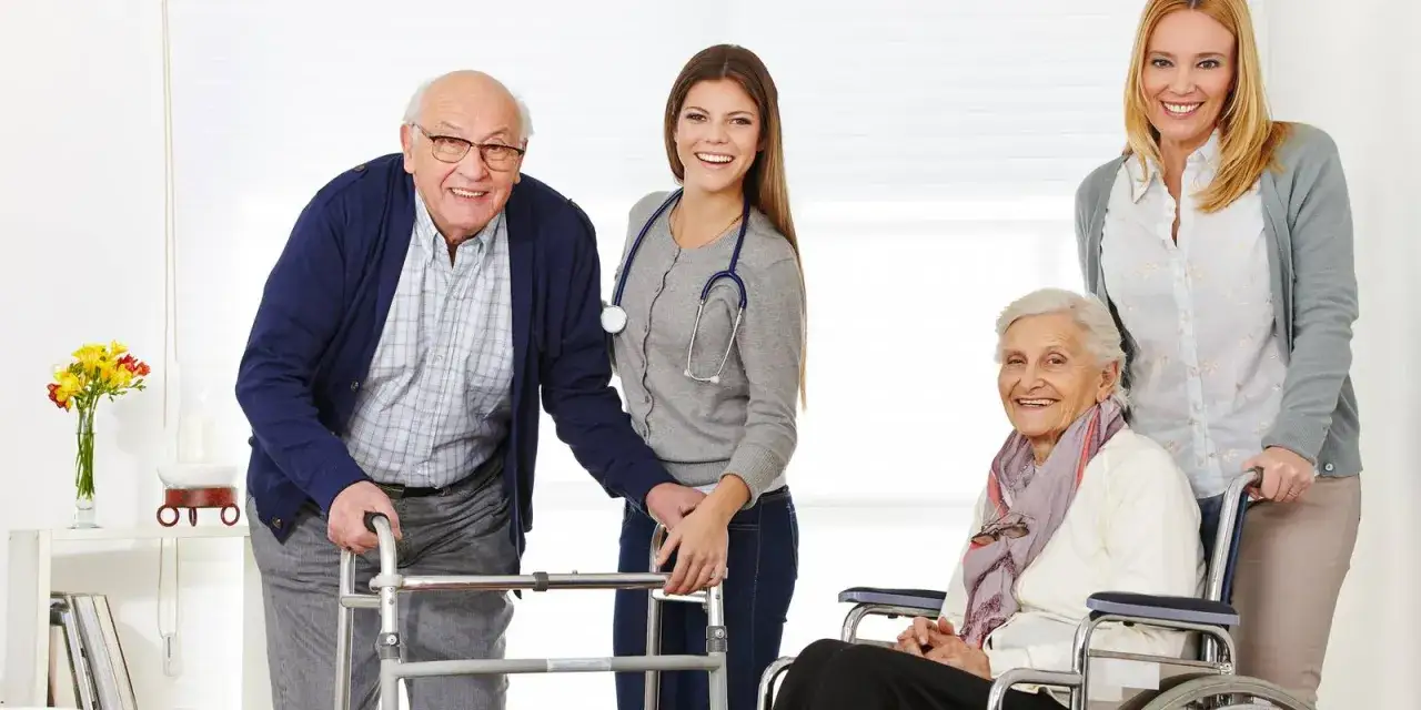 Un homme &acirc;g&eacute; utilise un d&eacute;ambulateur, une femme en fauteuil roulant est assist&eacute;e par deux soignantes. Une sc&egrave;ne de soutien et de soins, loin de l'hospitalisation.