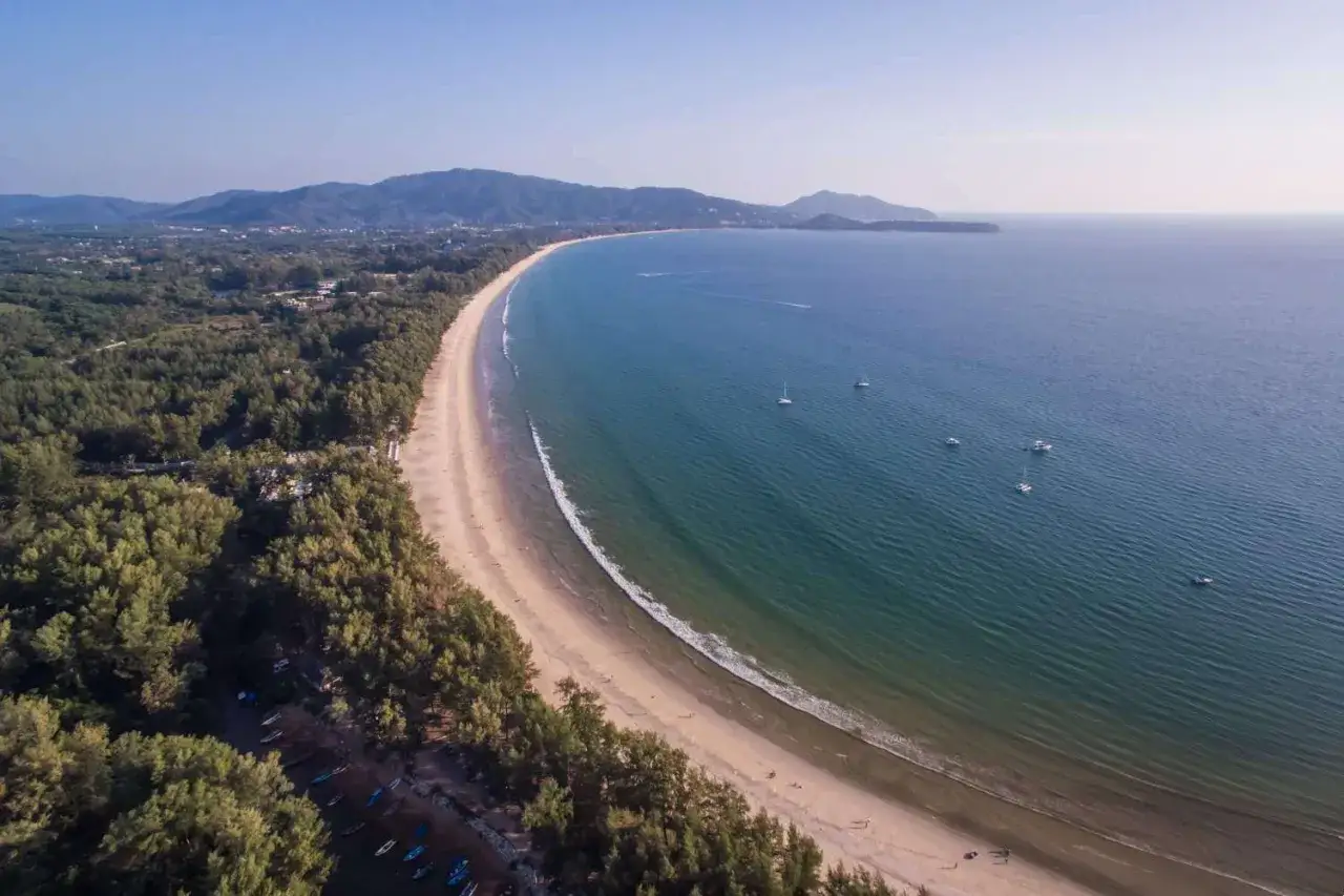 Vacker vy &ouml;ver Bang Tao Beach Phuket med turkost vatten, vita segelb&aring;tar och en l&aring;ng sandstrand kantad av gr&ouml;nska.