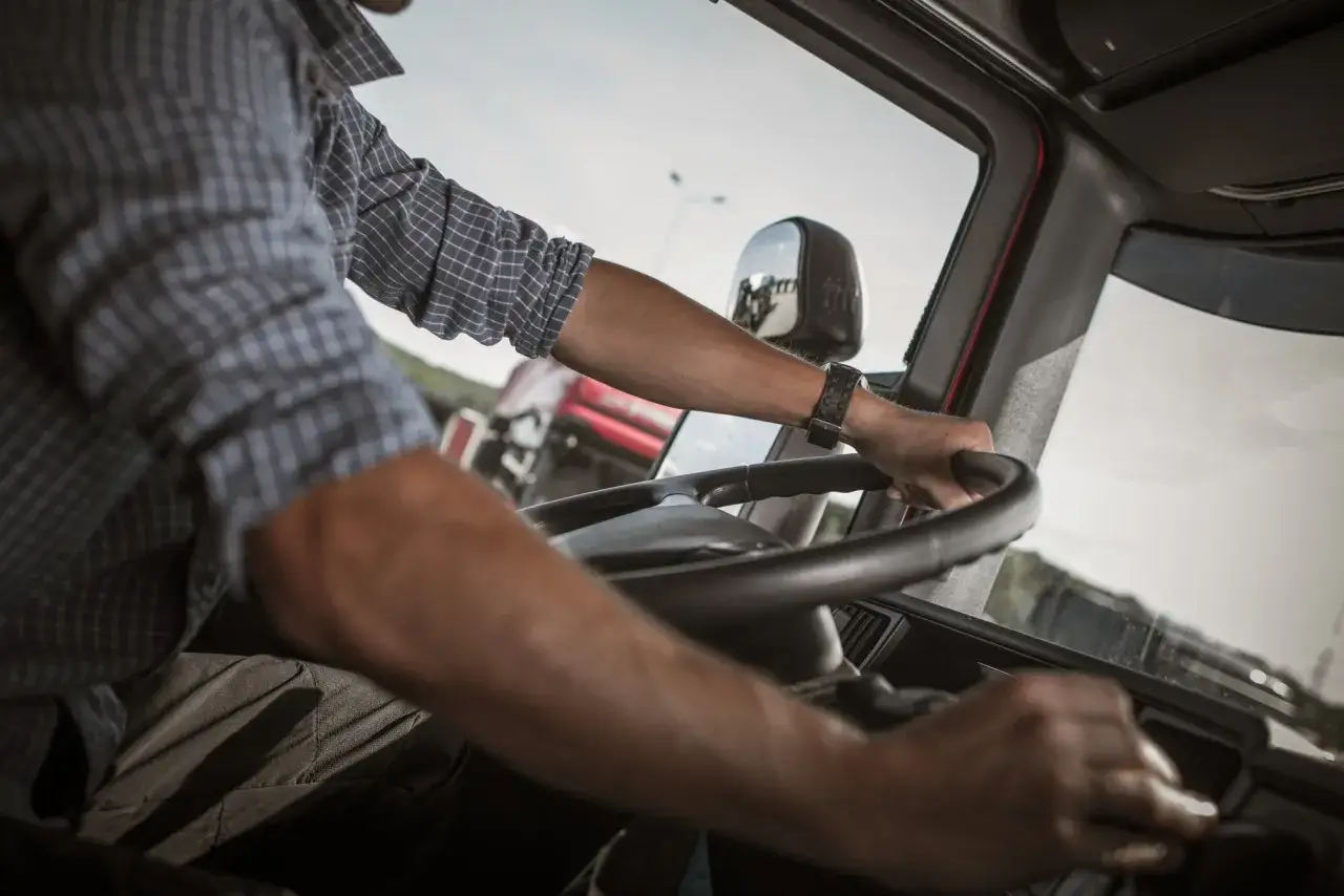 Conductor de camión al volante, asumiendo sus obligaciones. Se ve su mano en el volante y otra en la palanca de cambios.