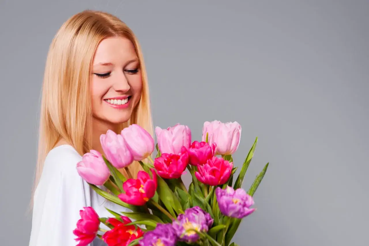 Uśmiechnięta kobieta z blond włosami trzyma bukiet kolorowych tulipan&oacute;w.