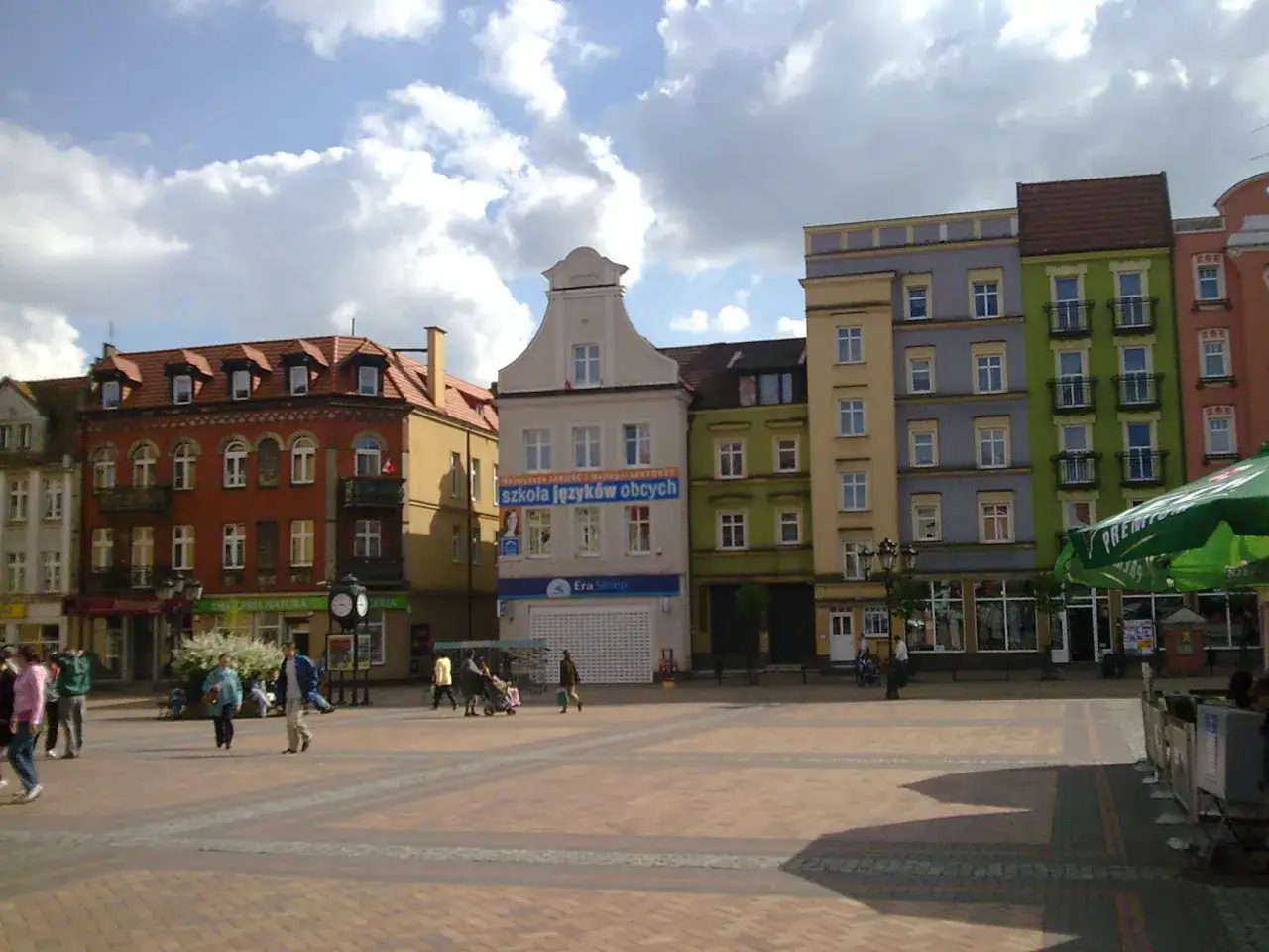 Rynek w Chojnicach z kolorowymi kamienicami, w tym ratusz. Ludzie spacerują po brukowanym placu.