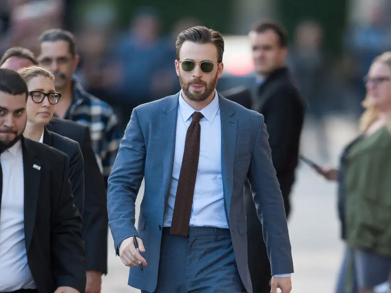 Mann in einem blauen Lounge Suit mit Sonnenbrille und Bart, der mit einer Gruppe von Menschen geht.
